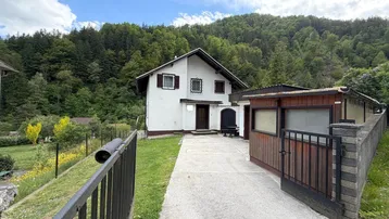 Weißes Einfamilienhaus mit braunem Dach und umgebender Natur, umgeben von Bäumen und Hügeln.