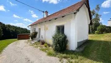 Kleines, älteres Haus mit rotem Ziegeldach und weißer Fassade, umgeben von viel Grün.