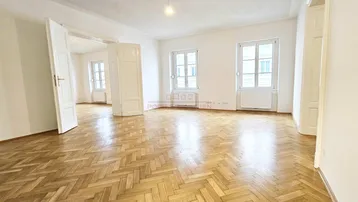 Geräumiges Wohnzimmer mit hellem Fischgrätparkett, weißen Wänden und hohen Fenstern, die viel Tageslicht hereinlassen.
