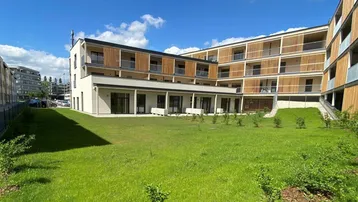 Modernes Wohngebäude mit Holzlamellenfassade und großer Rasenfläche im Vordergrund unter blauem Himmel.