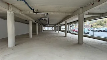 Geräumiger, unfertiger Gewerberaum mit Betonboden, Säulen und großen Fensterfronten zur Straße.