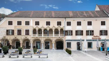 Historisches Gebäude mit aufwendiger Fassade, Arkaden und einem Café im Erdgeschoss an einem öffentlichen Platz.