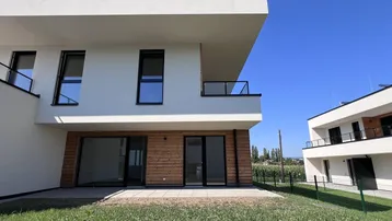Modernes Gebäude mit weißer Fassade, Holzverkleidung, Terrasse und Gartenbereich unter blauem Himmel.
