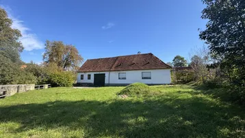 Ländliches Gebäude mit weißer Fassade, rotem Ziegeldach und großem Rasenbereich unter blauem Himmel.
