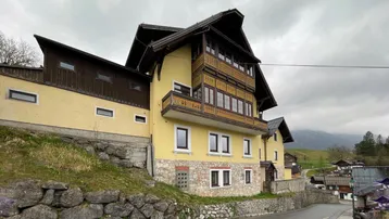 Traditionelles Gebäude mit gelber Fassade, Holzbalkonen und Steinsockel in alpiner Umgebung.