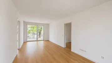 Helles, leeres Wohnzimmer mit Laminatboden und Zugang zu einem Balkon.