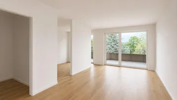 Heller, leerer Raum mit Holzboden und Zugang zu einem Balkon mit Blick ins Grüne.