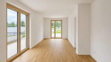 Helles, leeres Zimmer mit Holzboden und großen Fenstertüren, die zu einem Außenbereich führen.