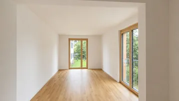 Helles, leeres Zimmer mit Holzboden und großen Fenstern, die einen Blick ins Grüne bieten.