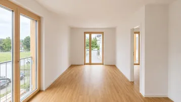 Heller, leerer Raum mit Holzboden und großen Fenstern, die viel Tageslicht hereinlassen.