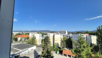 Weitblick über Wohngebäude und grüne Umgebung mit Bergen am Horizont unter blauem Himmel.