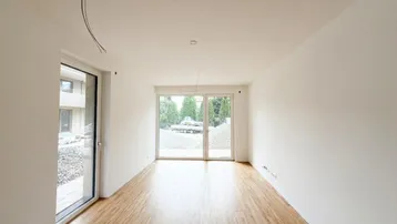 Helles, leeres Zimmer mit Holzboden und großen Fenstern, die viel Tageslicht hereinlassen.