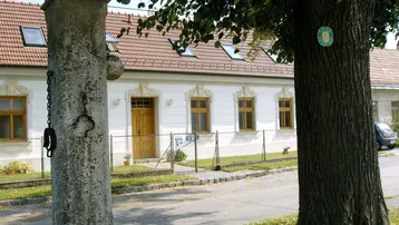 Traditionelles Gebäude mit Satteldach und Dachfenstern, umgeben von Bäumen und einem Zaun.