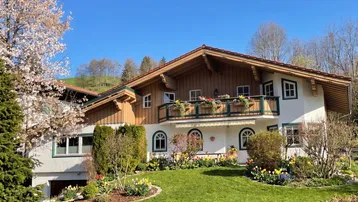 Traditionelles Alpenhaus mit gepflegtem Garten, blühenden Bäumen und Bergblick unter blauem Himmel.