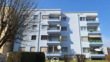 Mehrfamilienhaus mit hellblauer Fassade, mehreren Balkonen und gepflegter Grünanlage unter blauem Himmel.