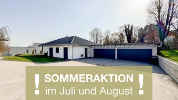 Modernes weißes Haus mit dunklem Satteldach und einer Doppelgarage, umgeben von gepflegten Grünflächen und einer Auffahrt.