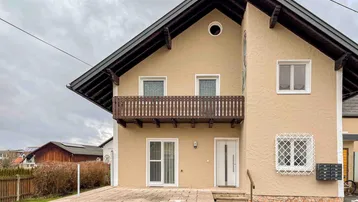Vorderansicht eines Einfamilienhauses mit Balkon, Terrasse und beiger Fassade unter bewölktem Himmel.