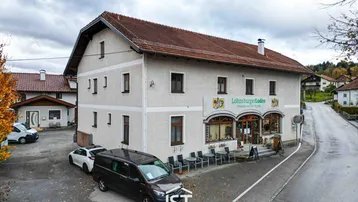 Gebäudeansicht mit Ladenlokal im Erdgeschoss und Wohnungen darüber, umgeben von ländlicher Umgebung.