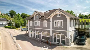 Luftaufnahme eines grauen Gebäudes mit weißer Fassadenverzierung und Satteldach an einer Straße.