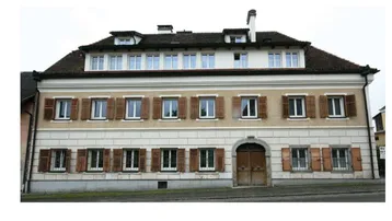 Klassisches Gebäude mit mehreren Fenstern und Holzläden, umgeben von einer hellen Fassade.