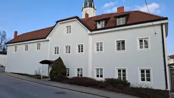 Historisches Gebäude mit heller Fassade und rotem Ziegeldach, im Hintergrund ein Kirchturm.
