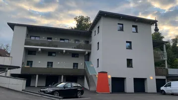 Modernes Mehrfamilienhaus mit mehreren Balkonen, Garagen und Außenparkplätzen unter bewölktem Himmel.