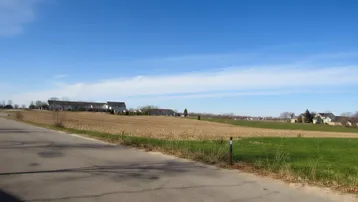 Weitläufige ländliche Landschaft mit Feldern und vereinzelten Häusern am Horizont unter einem strahlend blauen Himmel.