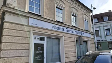 Fassade eines älteren Gebäudes mit Geschäftsräumen im Erdgeschoss und dekorativen Fenstern mit Rollläden.