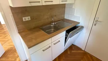 Kompakte Küche mit Spüle, Geschirrspüler und weißen Oberschränken über Holzoptik-Arbeitsplatte.