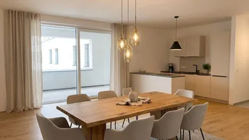 Heller offener Wohn-Essbereich mit Holztisch, modernen Pendelleuchten und Zugang zu einem Balkon.