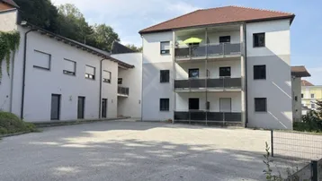 Mehrfamilienhaus mit mehreren Balkonen und großem Schotterparkplatz davor, umgeben von grüner Hügellandschaft.