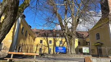 Historische gelbe Gebäude mit braunen Dächern, umgeben von hohen, kahlen Bäumen und einem schmiedeeisernen Zaun.