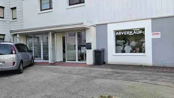 Außenansicht eines Mehrfamilienhauses mit einem Ladenlokal im Erdgeschoss und Parkplätzen davor.