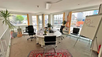 Heller Büroraum mit großen Fenstern, Teppichboden und einem Konferenztisch, ideal für Besprechungen.