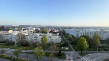 Weitläufiger Blick über eine Stadtlandschaft mit Wohngebäuden, Straßen und Grünflächen unter blauem Himmel.