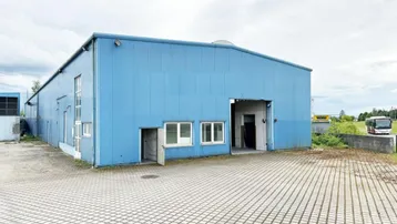 Außenansicht eines großen blauen Industriegebäudes mit gepflastertem Vorplatz und Zufahrt.