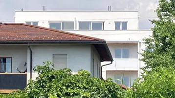 Neubau-Wohngebäude mit weißer Fassade, teilweise verdeckt von Bäumen und einem anderen Dach.