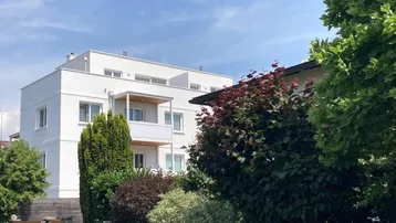 Modernes weißes Mehrfamilienhaus mit Balkonen, umgeben von üppiger grüner Bepflanzung unter blauem Himmel.