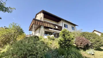 Hausfassade mit Balkon und umgebender Natur unter blauem Himmel.