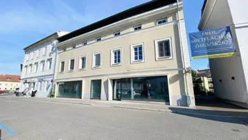 Gebäudeansicht von außen mit großen Schaufenstern im Erdgeschoss und einem Banner für freie Mietflächen.