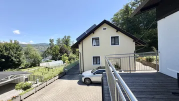 Ansicht des Hauses mit gepflasterter Auffahrt, Gartenbereich und Blick auf den See im Hintergrund.
