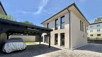 Modernes Haus mit beigefarbener Fassade und einem Carport für zwei Fahrzeuge auf gepflastertem Vorplatz.
