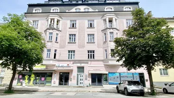 Mehrstöckiges Gebäude mit rosa Fassade, Geschäften im Erdgeschoss und Balkonen an einer belebten Straße.