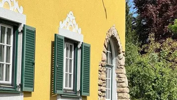 Historische Fassade mit gelbem Putz, grünen Fensterläden und rustikalem Steinsockel unter blauem Himmel.