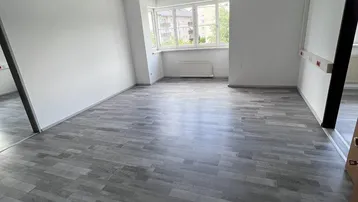 Geräumiger, leerer Büroraum mit grauem Laminatboden und großen Fenstern, die viel Tageslicht hereinlassen.