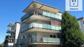 Modernes Mehrfamilienhaus mit mehreren Balkonen und Glasbrüstungen unter blauem Himmel.