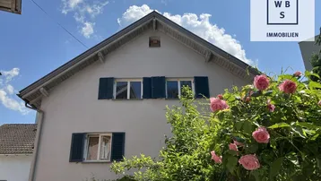 Charmantes Haus mit Satteldach, grünen Fensterläden und blühenden Rosen im Vorgarten.