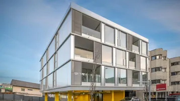 Modernes Gebäude mit Glasfassade und gelben Stützen, umgeben von einer städtischen Umgebung.