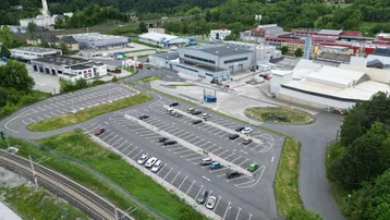 Luftaufnahme eines Industriegebiets mit großem Parkplatz, umgeben von grünen Hügeln und Gebäuden.