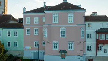 Historisches Gebäude mit rosa und hellgrüner Fassade, traditionellen Fenstern und Satteldach in einer städtischen Umgebung.